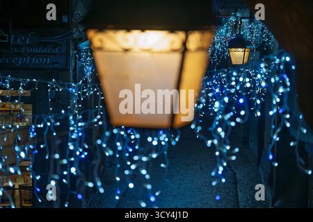 Blau-weiße Lichterketten beleuchten eine charmante Gasse in Mont Saint-Michel, Frankreich, und schaffen eine festliche Atmosphäre rund um ein historisches Hotel. Stockfoto