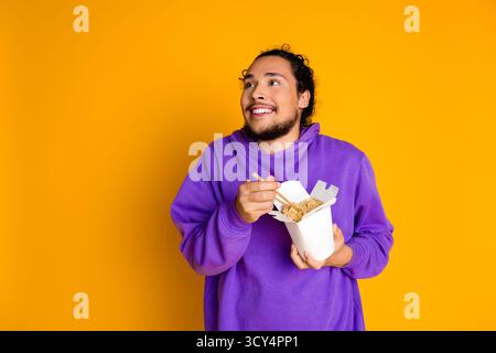 Junger Mann in violettem Hoodie, der Nudeln genießt, während er vor leuchtendem gelbem Hintergrund lächelt Stockfoto