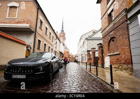 Tarnow, Polen - 10. Oktober 2025: Ein stilvoller schwarzer Maserati Levante parkt entlang einer nassen Kopfsteinpflasterstraße in Tarnow in der Nähe von Backsteinhäusern. Stockfoto