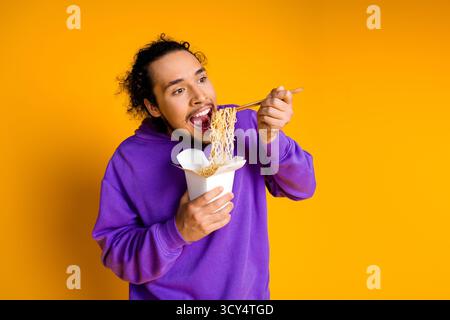 Junger Mann mit violettem Hoodie, der asiatische Nudeln mit Essstäbchen auf leuchtendem gelbem Hintergrund genießt und den lässigen Lebensstil darstellt Stockfoto