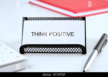 Ein kleiner Halter mit einer Karte mit der Aufschrift „Think positive“ sitzt auf einem sauberen Schreibtisch. In der Nähe bieten ein glänzender Stift und ein hellrotes Notizblock einen kreativen Arbeitsplatz Stockfoto