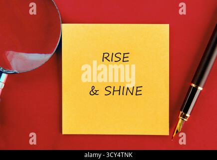 Eine leuchtend gelbe Haftnotiz mit den Worten „Rise and Shine“, begleitet von einem Stift und einer Lupe, liegt auf einer kräftigen roten Oberfläche und ist einladend Stockfoto