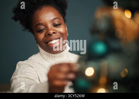 Schwarze junge Erwachsene Frau lächelt, während sie den Weihnachtsbaum mit Ornamenten dekoriert, einen Rollkragenpullover trägt und sich auf festliche Feiertage im Innenbereich konzentriert Stockfoto