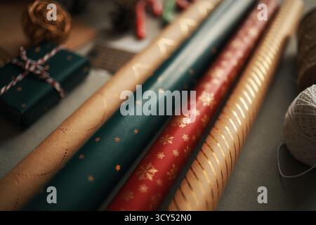 Nahaufnahme mit Rollen Weihnachtspapier mit festlichen Mustern und Geschenkbox in der Nähe, darunter goldene Sterne, Schneeflocken und dekorative Garne für Weihnachtsgeschenke Stockfoto