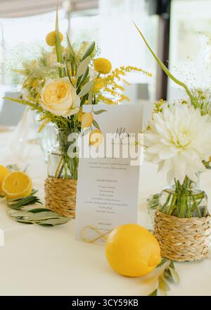 Wunderschöne Tischdekoration mit eleganter Menükarte umgeben von leuchtend gelben Rosen und Zitronen. Natürliche Blumenarrangements sorgen für eine erfrischende Atmosphäre Stockfoto