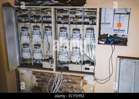 Eine freiliegende elektrische Schalttafel in einem Wohnhaus, die mehrere Meter, verwickelte Drähte und sichtbare Wandschäden zeigt Stockfoto