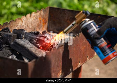 Close-up of a hand in a glove, using a butane torch to quickly start fire and ignite black charcoal briquettes in a rusty metal BBQ grill Stockfoto