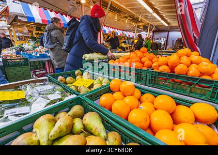Ein Käufer wählt frische Produkte auf einem lebhaften Markt im Freien, umgeben von Kisten mit Orangen und Birnen Stockfoto