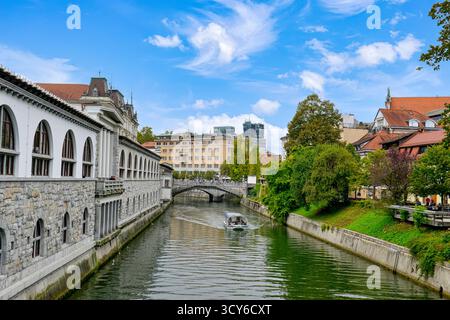 Blick auf die Touristenboote auf der Ljubljanica, Cafés und andere Gebäude entlang der Uferpromenade in Ljubljana, Slowenien Stockfoto