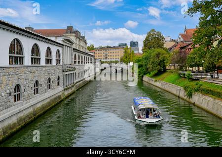 Blick auf die Touristenboote auf der Ljubljanica, Cafés und andere Gebäude entlang der Uferpromenade in Ljubljana, Slowenien Stockfoto