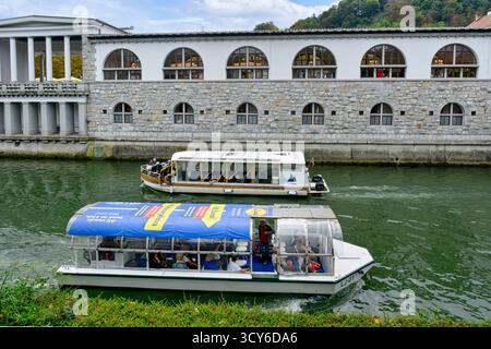Blick auf die Touristenboote auf der Ljubljanica, Cafés und andere Gebäude entlang der Uferpromenade in Ljubljana, Slowenien Stockfoto