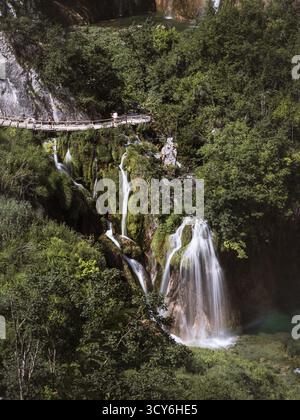 Ein detaillierter Blick auf Wasserfälle, die über moosige Felsen stürzen, umgeben von dichtem Grün, mit Touristen, die auf einer hölzernen Promenade über den Kaskaden i spazieren Stockfoto