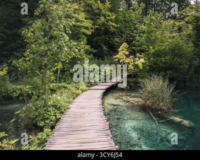 Ein geschwungener Holzsteg schlängelt sich durch üppiges Grün und über kristallklares türkisfarbenes Wasser in den Plitvicer Seen, Kroatien, und schafft eine ruhige Landschaft Stockfoto