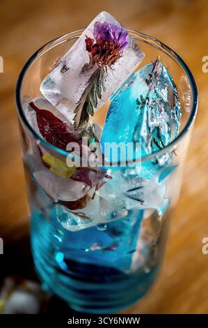 Glas gefüllt mit Eiswürfeln mit eingebetteten Blumen, in leuchtend blauer Flüssigkeit. Stockfoto