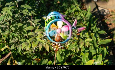 Ein skurriles Regenbogenfarbiges, vogelförmiges Windrad steht still im üppigen Gartengrün und verbindet verspielte Dekoration mit natürlicher Schönheit. Stockfoto