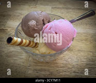 Drei Kugeln Eis – Erdbeere, Schokolade und Vanille – serviert in einer Dessertschüssel aus Glas mit einem Waffelstab. Erfrischendes Sommerdessert Stockfoto