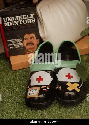 Nahaufnahme von Hochglanz-Schwesternclogs auf Kunstrasen neben einem Buch über „unwahrscheinliche Mentoren“ mit Stalin. Dunkel satirisch und konzeptuell. Stockfoto