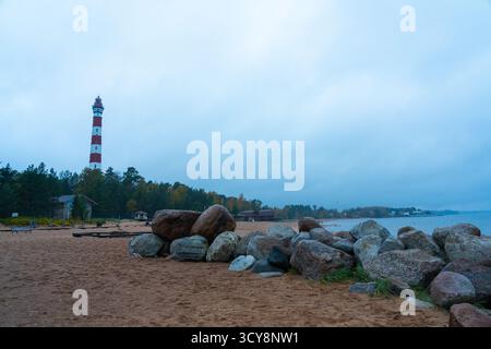 Ein hoher, rot-weiß gestreifter Leuchtturm am Ufer eines breiten Sandstrandes, eingerahmt von einer Linie aus schützenden Felsbrocken Stockfoto