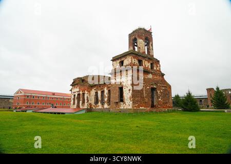 Shlisselburg, Russland - 13. Mai 2024: Die stark beschädigten Backsteinruinen der ehemaligen Gefängniskirche stehen auf einem grünen Rasen neben einem restaurierten roten Backsteinbund Stockfoto