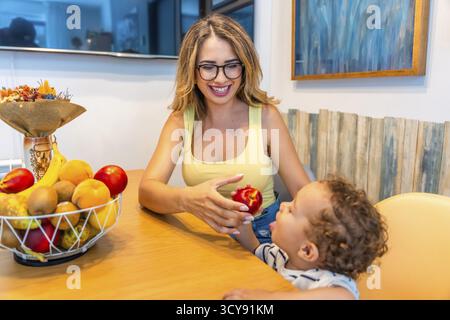 Liebevolle Mutter, die ihrem kleinen Sohn einen frischen Pfirsich schenkt, während sie am Küchentisch sitzt und gesunde Essgewohnheiten fördert Stockfoto