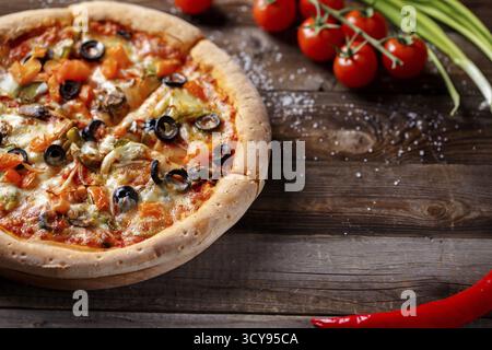 Vegetarische Pizza mit Pilzen und Oliven auf einem alten Holztisch und frischen Tomaten, grünen Zwiebeln und Chili-Pfeffer im Hintergrund Stockfoto
