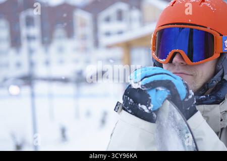 Mann-Snowboarder tragen orange Helm, graue Jacke, schwarze und blaue Handschuhe stehend mit snowboard Stockfoto
