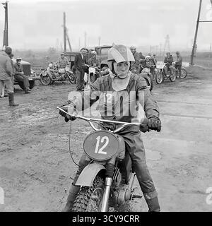Ein dynamisches Archivfoto aus den 1970er Jahren fängt die düstere Atmosphäre eines Motocross-Rennens in Sloviansk, der ukrainischen SSR, ein. Fahrer mit Vintage-Helmen und Schutzausrüstung sitzen auf ihren leistungsstarken Motorrädern aus der Sowjetzeit (wahrscheinlich CZ- oder Izh-Modelle) auf einer schlammigen Feldpiste, bereit für den Wettkampf. Zuschauer treffen sich in der Nähe, was die Spannung des lokalen Motorsport-Events noch verstärkt. Dieses Bild zeigt die Leidenschaft für Sport und Gemeindeversammlungen in einem friedlichen Donbass vor dem Krieg Stockfoto