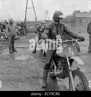 Ein dynamisches Archivfoto aus den 1970er Jahren fängt die düstere Atmosphäre eines Motocross-Rennens in Sloviansk, der ukrainischen SSR, ein. Fahrer mit Vintage-Helmen und Schutzausrüstung sitzen auf ihren leistungsstarken Motorrädern aus der Sowjetzeit (wahrscheinlich CZ- oder Izh-Modelle) auf einer schlammigen Feldpiste, bereit für den Wettkampf. Zuschauer treffen sich in der Nähe, was die Spannung des lokalen Motorsport-Events noch verstärkt. Dieses Bild zeigt die Leidenschaft für Sport und Gemeindeversammlungen in einem friedlichen Donbass vor dem Krieg Stockfoto