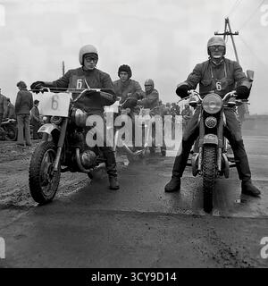 Ein dynamisches Archivfoto aus den 1970er Jahren fängt die düstere Atmosphäre eines Motocross-Rennens in Sloviansk, der ukrainischen SSR, ein. Fahrer mit Vintage-Helmen und Schutzausrüstung sitzen auf ihren leistungsstarken Motorrädern aus der Sowjetzeit (wahrscheinlich CZ- oder Izh-Modelle) auf einer schlammigen Feldpiste, bereit für den Wettkampf. Zuschauer treffen sich in der Nähe, was die Spannung des lokalen Motorsport-Events noch verstärkt. Dieses Bild zeigt die Leidenschaft für Sport und Gemeindeversammlungen in einem friedlichen Donbass vor dem Krieg Stockfoto