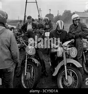 Ein dynamisches Archivfoto aus den 1970er Jahren fängt die düstere Atmosphäre eines Motocross-Rennens in Sloviansk, der ukrainischen SSR, ein. Fahrer mit Vintage-Helmen und Schutzausrüstung sitzen auf ihren leistungsstarken Motorrädern aus der Sowjetzeit (wahrscheinlich CZ- oder Izh-Modelle) auf einer schlammigen Feldpiste, bereit für den Wettkampf. Zuschauer treffen sich in der Nähe, was die Spannung des lokalen Motorsport-Events noch verstärkt. Dieses Bild zeigt die Leidenschaft für Sport und Gemeindeversammlungen in einem friedlichen Donbass vor dem Krieg Stockfoto