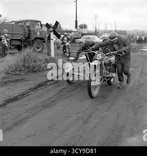 Ein dynamisches Archivfoto aus den 1970er Jahren fängt die düstere Atmosphäre eines Motocross-Rennens in Sloviansk, der ukrainischen SSR, ein. Fahrer mit Vintage-Helmen und Schutzausrüstung sitzen auf ihren leistungsstarken Motorrädern aus der Sowjetzeit (wahrscheinlich CZ- oder Izh-Modelle) auf einer schlammigen Feldpiste, bereit für den Wettkampf. Zuschauer treffen sich in der Nähe, was die Spannung des lokalen Motorsport-Events noch verstärkt. Dieses Bild zeigt die Leidenschaft für Sport und Gemeindeversammlungen in einem friedlichen Donbass vor dem Krieg Stockfoto