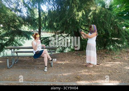 Zwei Frauen genießen einen sonnigen Nachmittag in einem Park, während eine Fotos von der anderen macht, während sie auf einem Picknicktisch sitzen, umgeben von üppigen grünen Bäumen und Stockfoto