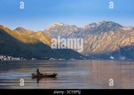 Goldener Sonnenaufgang über der Bucht von Kotor, Montenegro, mit einem kleinen Boot auf ruhigem Wasser und majestätischen Bergen, die vom Morgenlicht beleuchtet werden. Stockfoto