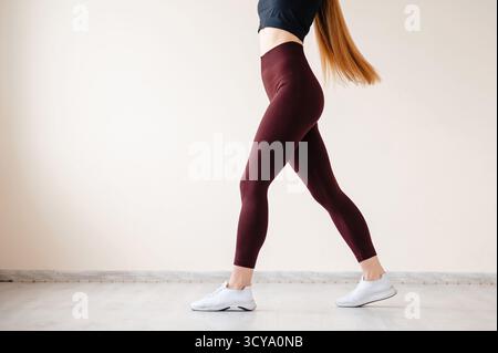 Eine Frau in burgunderroten Leggings und einem schwarzen Crop Top geht anmutig, zeigt Fitness und Bewegung, mit einem neutralen Hintergrund Stockfoto