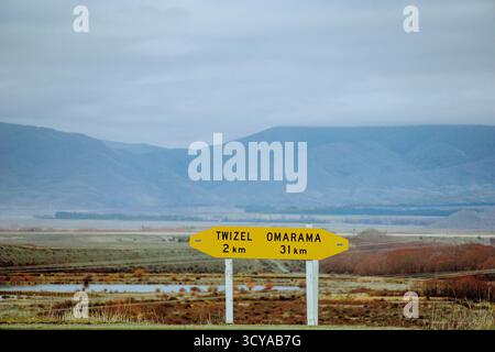 Ein gelbes Straßenschild zeigt die Entfernungen zu Twizel und Omarama an, die sich vor einer verhangenen Bergkette und einer trockenen Landschaft befinden. Stockfoto