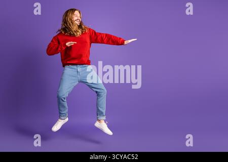 Junger Mann springt in lila Studio, trägt ein rotes Hemd, lächelt und hat Spaß Stockfoto