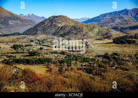 Ein Blick aus einem hohen Winkel fängt eine Tallandschaft mit einer Autobahn, verstreuten Häusern, Feldern und einem sich windenden Fluss ein, der von Hügeln und schneebedeckten Bergen eingerahmt wird. Stockfoto
