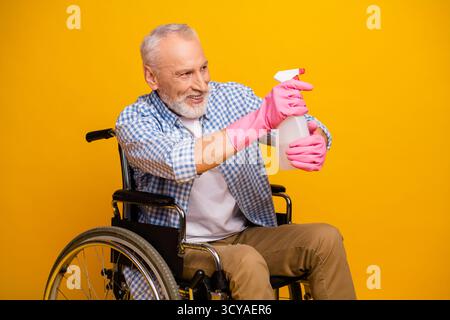 Opa im Rollstuhl sprüht mit pinkfarbenen Handschuhen vor hellgelbem Hintergrund Reiniger und zeigt dabei Resilienz und Humor Stockfoto