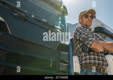 Ein Lkw-Fahrer steht selbstbewusst neben seinem Lkw, trägt eine Sonnenbrille und ein kariertes Hemd bei hellem Sonnenlicht. Stockfoto