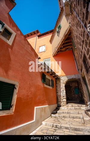 Rovinj, Kroatien - 14. August 2025: Eine sonnige Gasse mit Terrakotta- und Steinmauern, bunten Fensterläden und einem kleinen Bogen, der durch enge hinauf führt Stockfoto