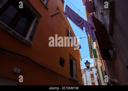 Rijeka, Kroatien - 14. August 2025: Wäscherei hängt über einer engen Gasse zwischen ockerfarbenen und pastellfarbenen Wänden, mit offenen Fensterläden und einer alten Straßenlaterne Stockfoto