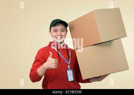 Glückliche Lieferperson in rotem Hoodie mit Paketboxen, die im Studiohintergrund die Daumen hochlegen Stockfoto