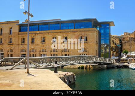 Bormla, Malta - 07.17.2025: Stilvolle Fußgängerbrücke aus Metall über ruhiges Wasser, die zu einem Café auf Treppen führt, neben einem Steingebäude mit Bogenfenstern Stockfoto