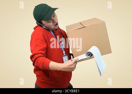 Ein geschäftiger Liefermann am Telefon mit großem Karton und Papierrolle in lässigem rotem Hoodie Stockfoto