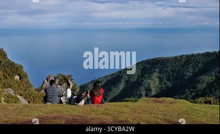 Drei Personen sitzen auf einer grasbewachsenen Klippe mit Blick auf das Meer und die bewaldeten Berge. Malerischer Blick auf die Küste mit felsigen Ausläufern, blauem Wasser und friedlicher Umgebung Stockfoto