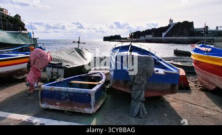Farbenfrohe Fischerboote auf Betonsteg am Meer, mit felsiger Küste und weißem Leuchtturm im Hintergrund. Friedliche maritime Szene unter teilweise Clou Stockfoto