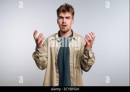Ein junger Mann steht in einer neutralen Umgebung und zeigt Überraschung und Verwirrung mit leicht erhobenen Armen. Stockfoto