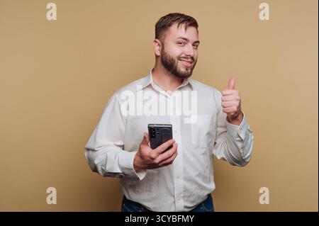 Ein Mann mit weißem Hemd steht vor einem beigefarbenen Hintergrund, hält ein Smartphone in der einen Hand und gibt mit der anderen einen Daumen hoch. Er lächelt Stockfoto