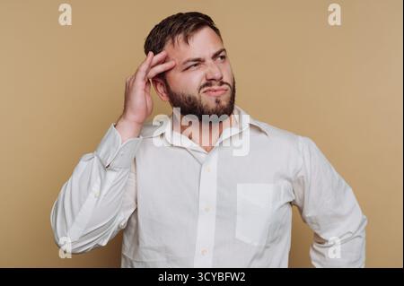 Ein Mann mit Bart sieht verwirrt aus, während er seinen Kopf in Gedanken berührt. Er trägt ein weißes Hemd und steht vor einem weichen beigen Hintergrund, was auf Dee hindeutet Stockfoto
