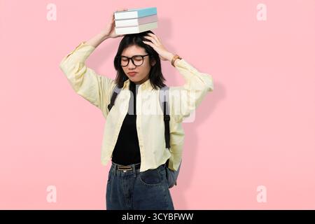 Junge Frau mit Rucksack, die einen Stapel Bücher auf dem Kopf vor rosa Hintergrund balanciert Stockfoto